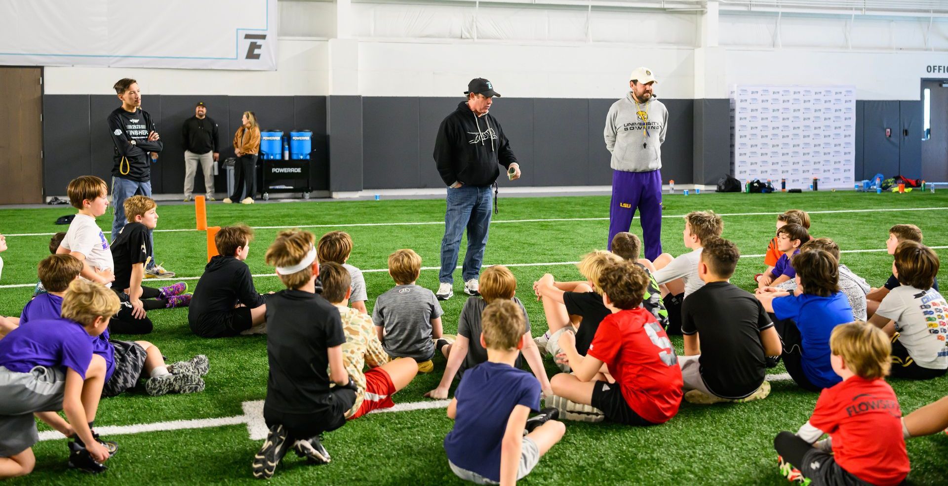 Children seated on indoor turf listening to two instructors at a sports facility