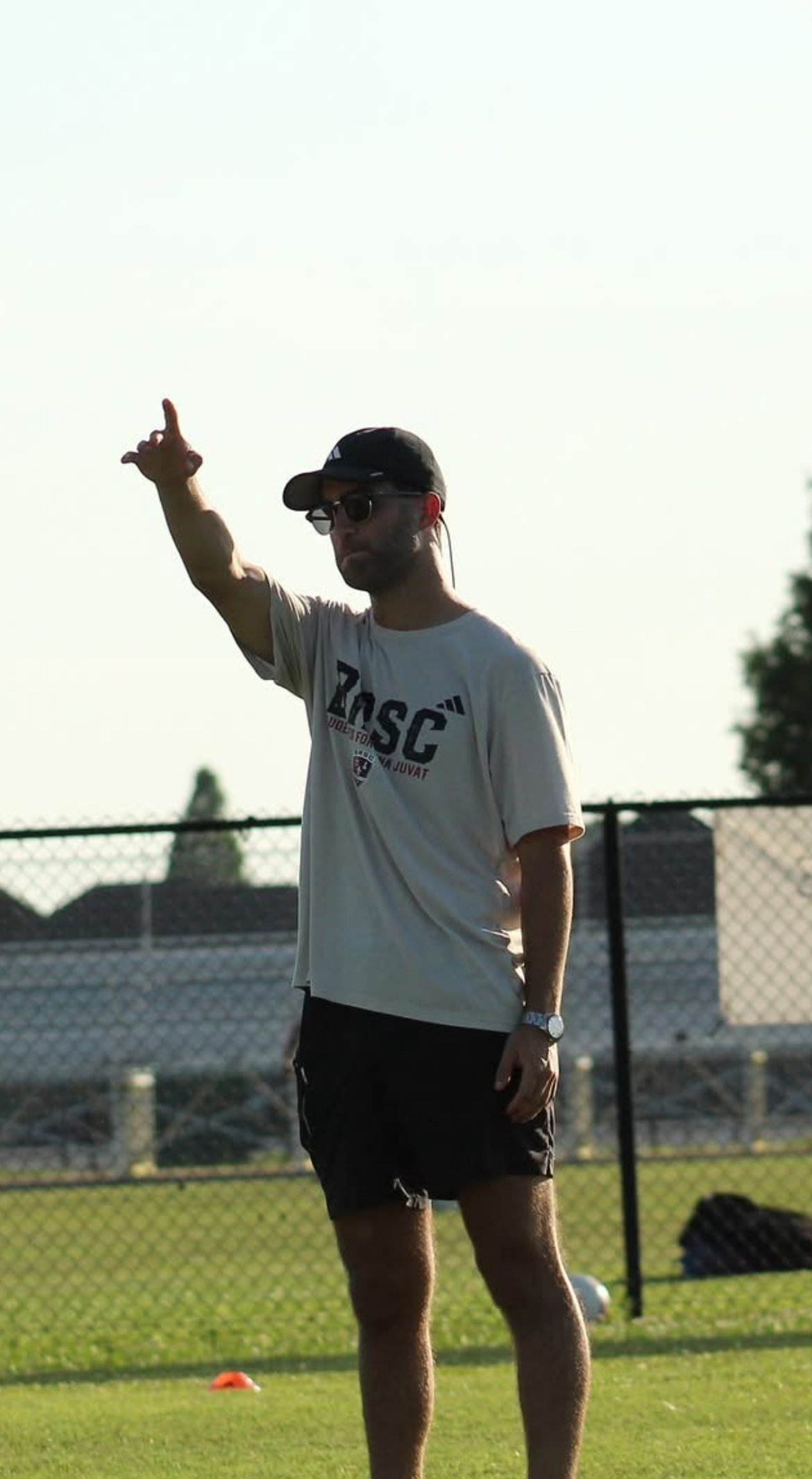 Person in a white shirt and black shorts stands on a sports field, pointing upward with one arm.