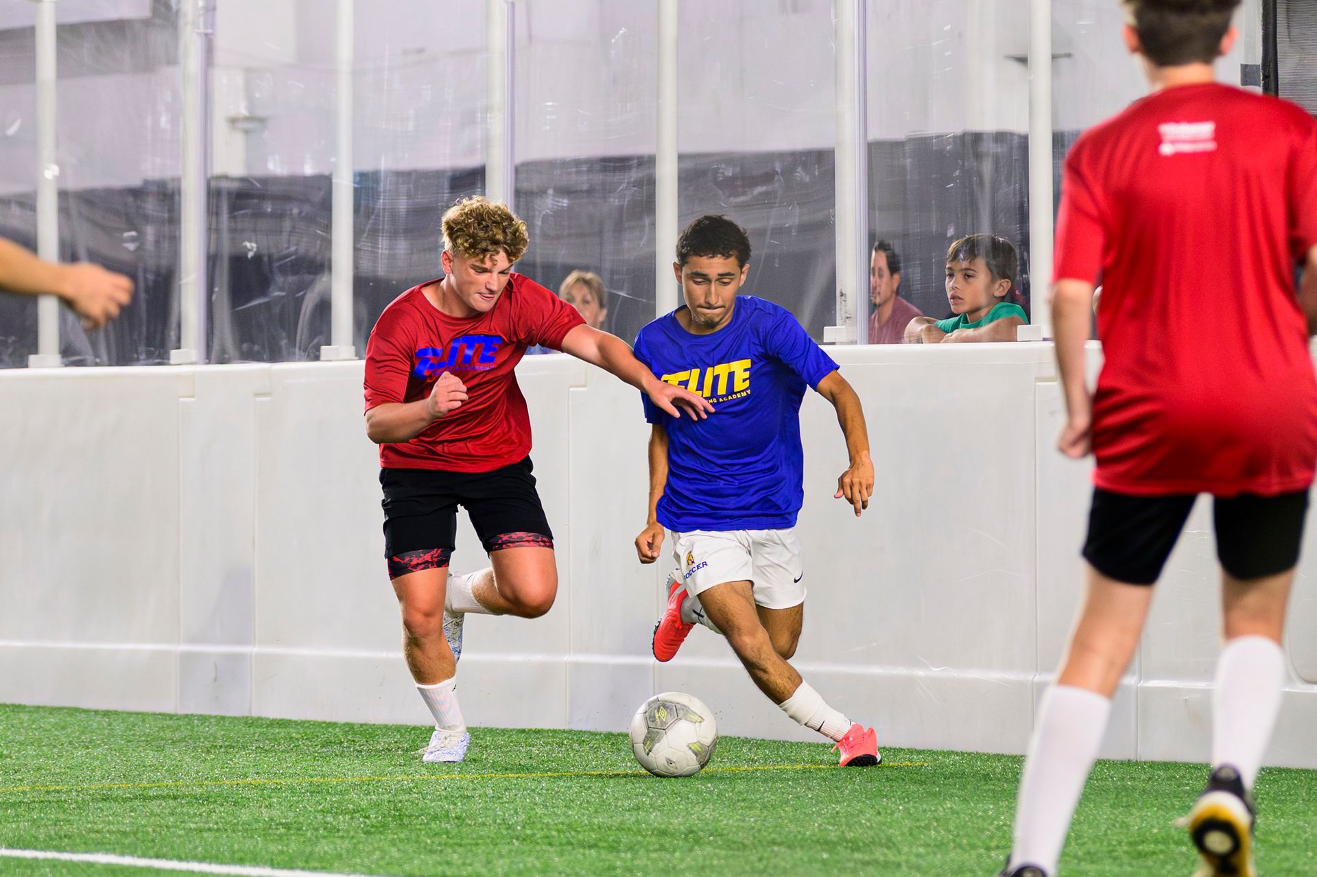Soccer players in red and blue jerseys battle for the ball on an indoor turf field