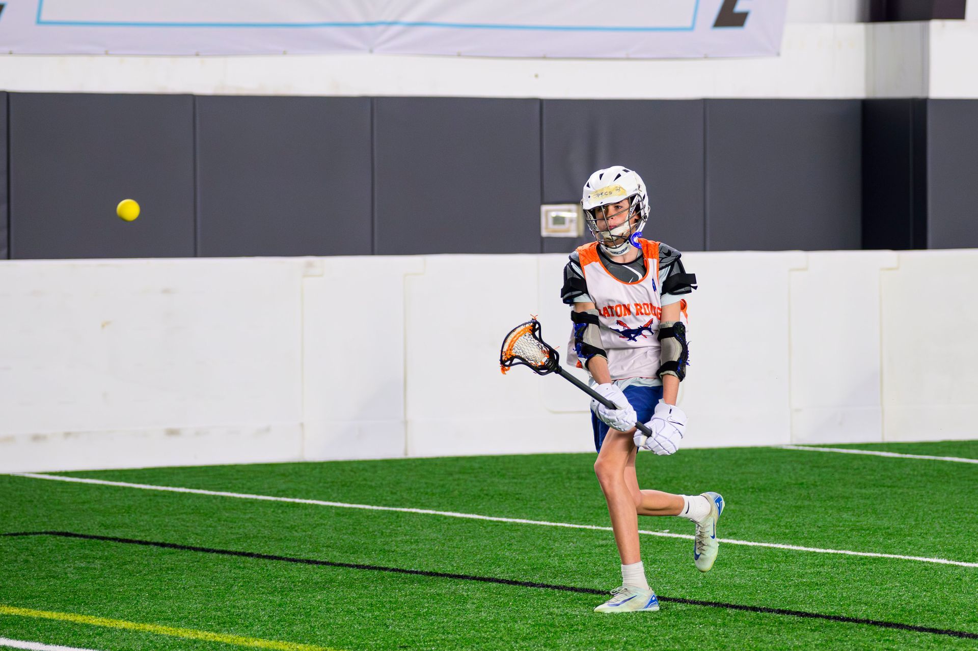 Lacrosse player in white and orange runs on indoor turf, holding a stick near a yellow ball.