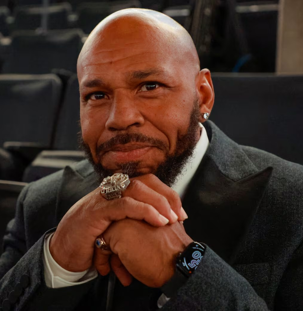 Close-up of a man in a dark suit, hands clasped, wearing a watch and ring, seated in an auditorium.
