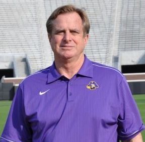 Man in purple polo shirt standing outdoors in front of a stadium seating area
