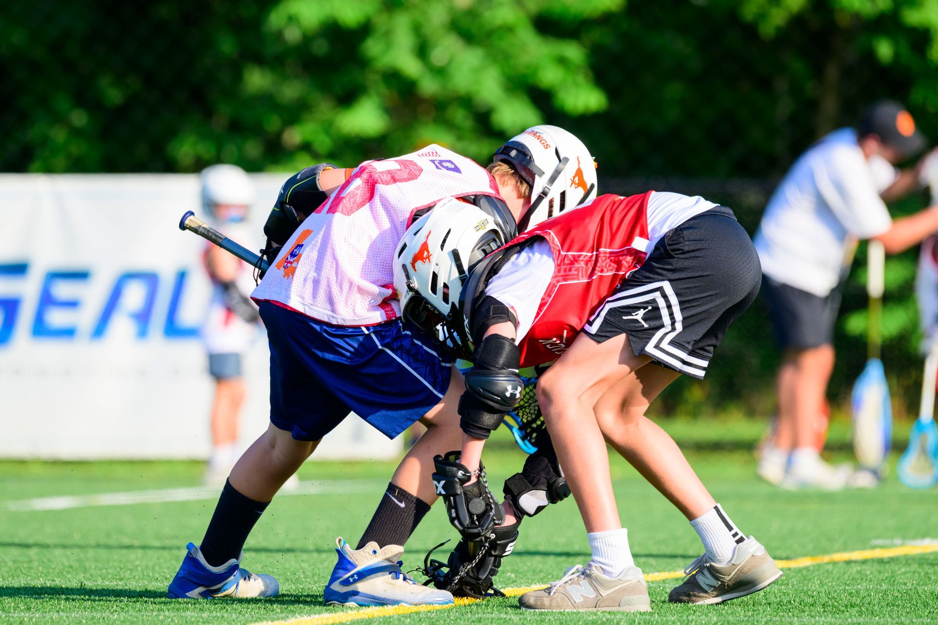 Two lacrosse players crouch low, fighting for the ball on a grassy field.