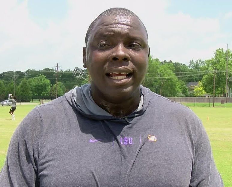 Man speaking outdoors on a sunny sports field, wearing a gray shirt and neck gaiter.
