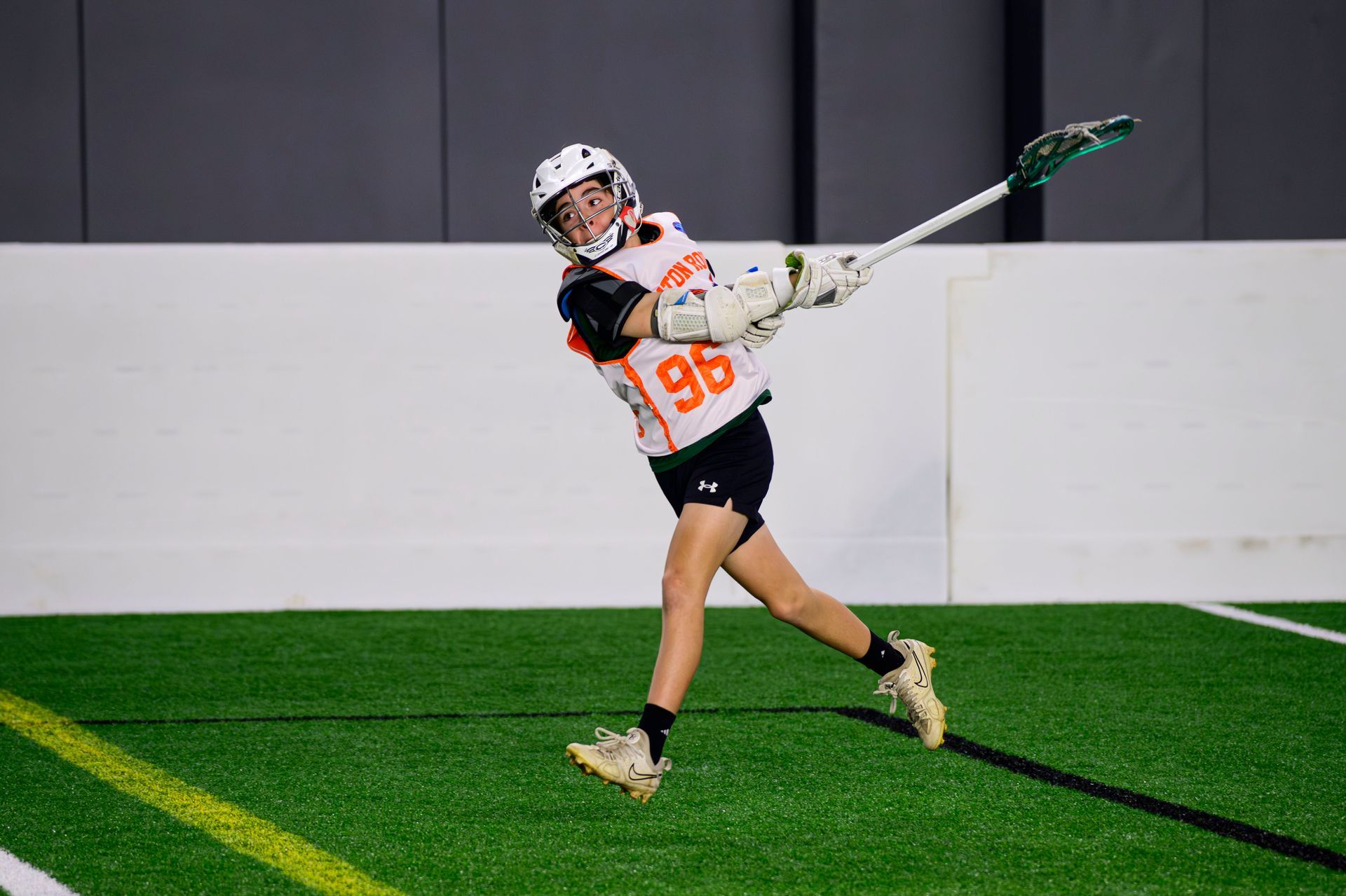 Lacrosse player in white and orange uniform swinging a stick on indoor turf field