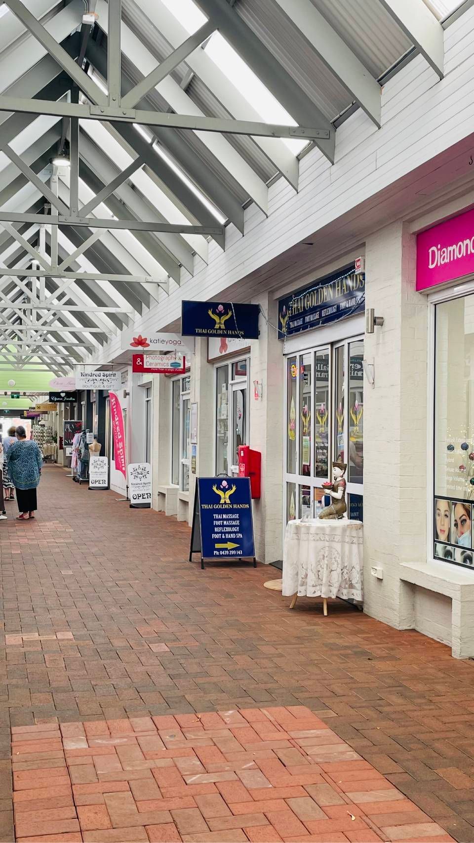 Covered shopping arcade with brick walkway and storefronts; a few people walk in the distance.