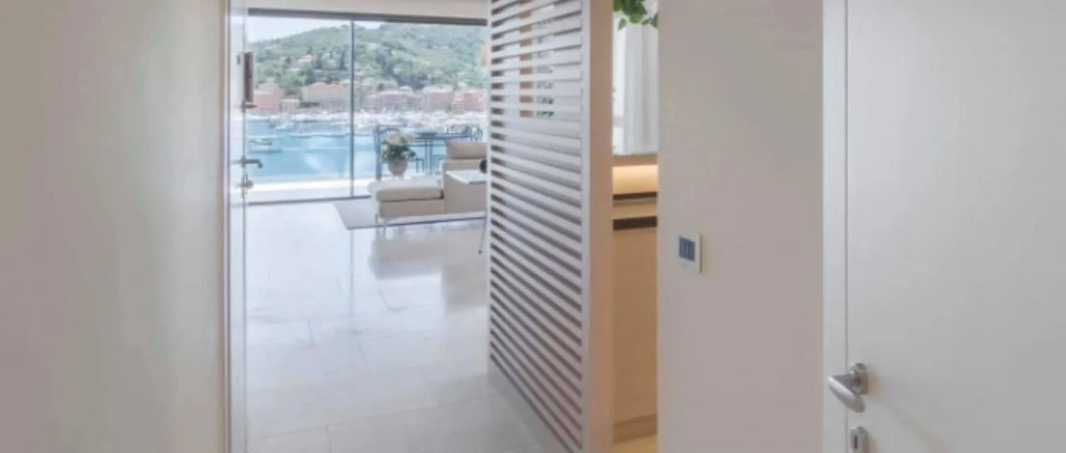 Luminoso corridoio che si apre su una porta scorrevole in vetro e su un balcone, con pareti bianche e pavimento piastrellato.