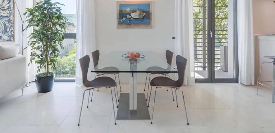 Sala da pranzo luminosa con tavolo in vetro, quattro sedie, pianta in vaso e opera d'arte sopra il tavolo.