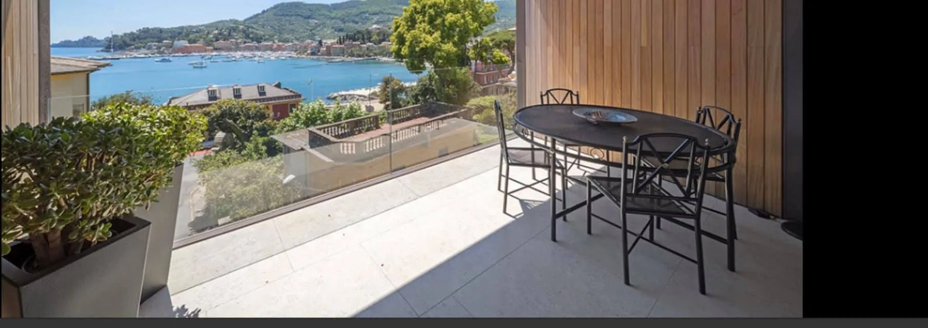 Balcone con tavolo e sedie neri che si affaccia su una soleggiata baia blu e sulle case sulla collina.