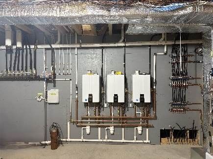Three white wall-mounted boilers with copper and white pipes, on a gray basement wall.