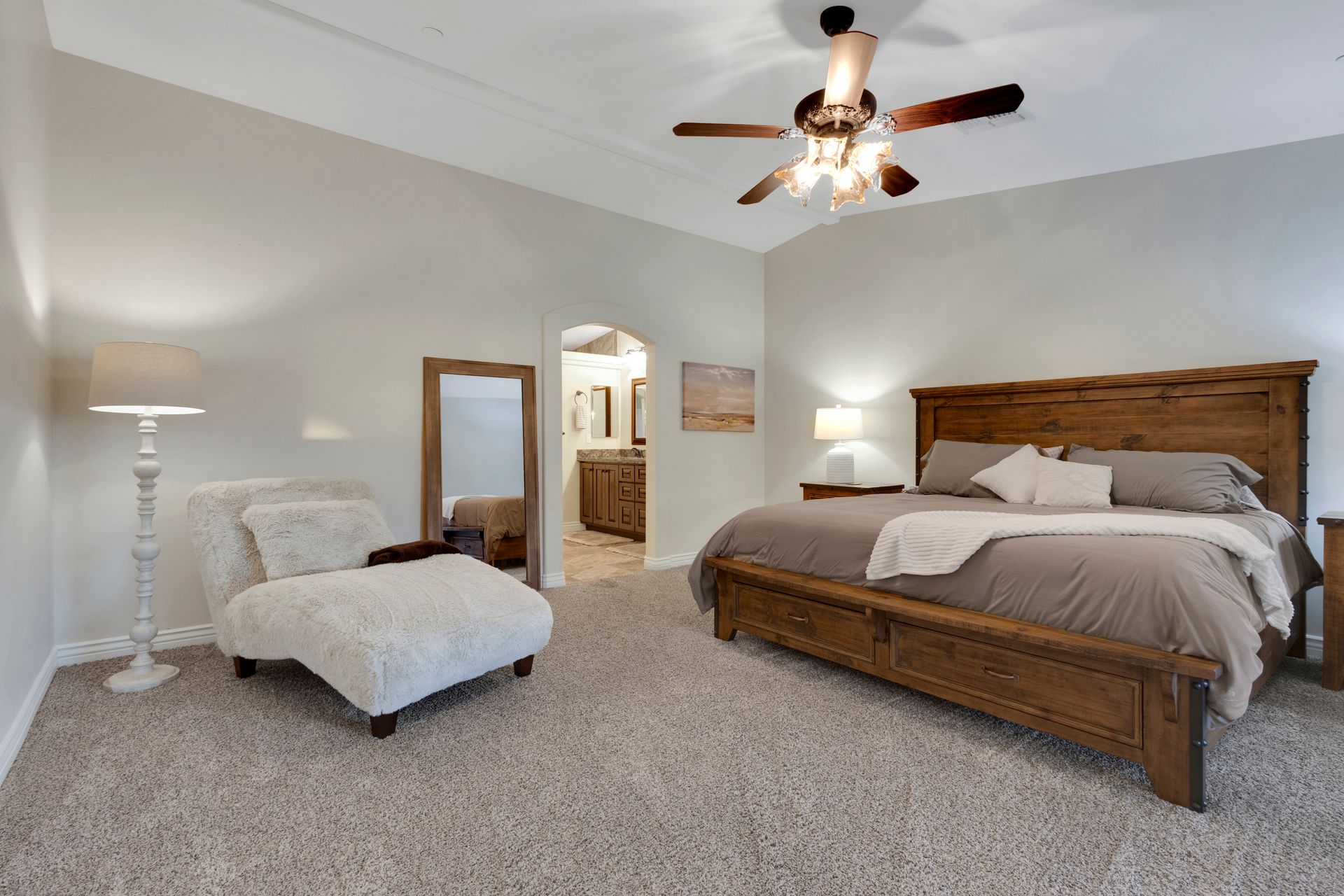 Bedroom with a wooden bed, sitting chair, and a mirror. Light-colored walls, carpet, and a ceiling fan.