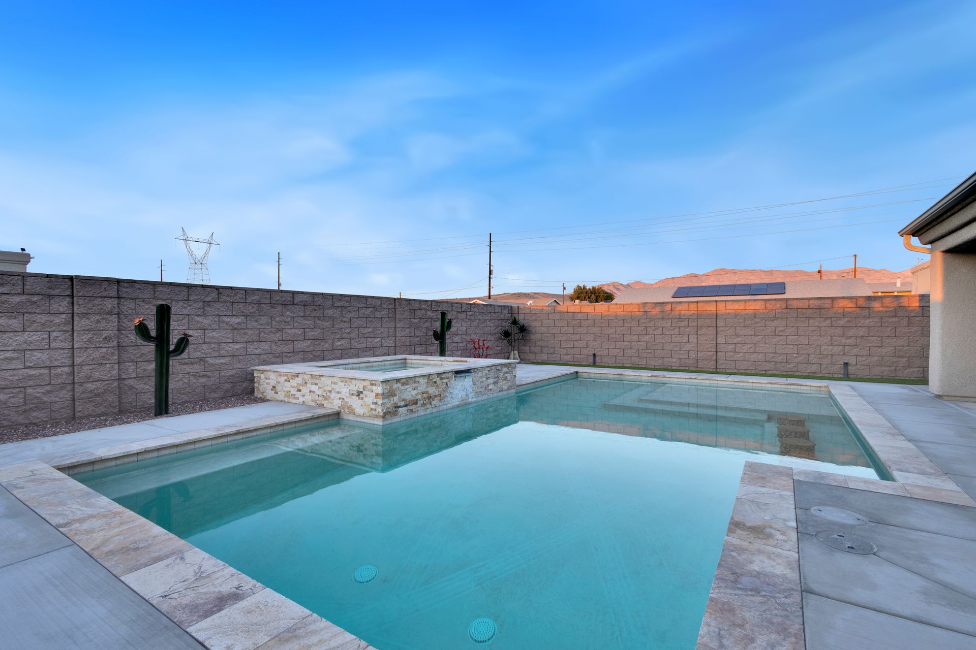 A backyard pool with a stone fire pit, cactus, and a brick wall under a blue sky.