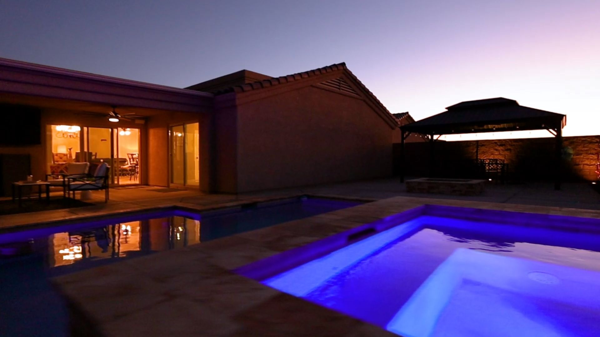 A pool with blue lights, a patio with furniture, and a gazebo are lit up at dusk.