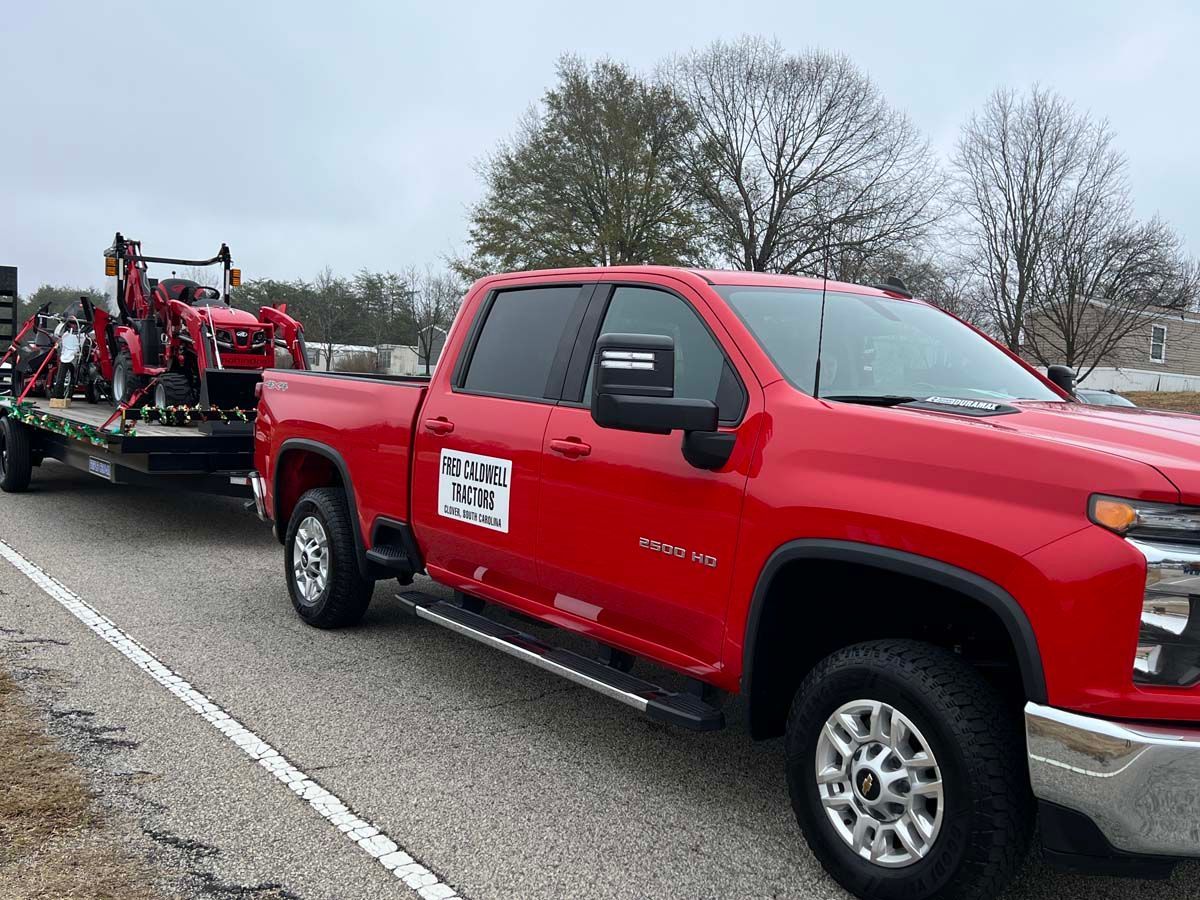 Red pickup truck towing a red farm machine on a trailer along a wet roadside