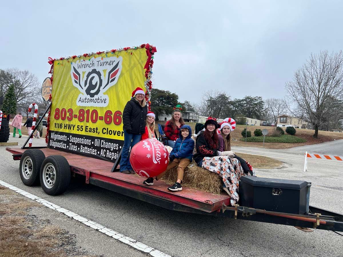 Decorated parade float with seated people, a large red balloon, and a sign on a trailer on a suburban street.