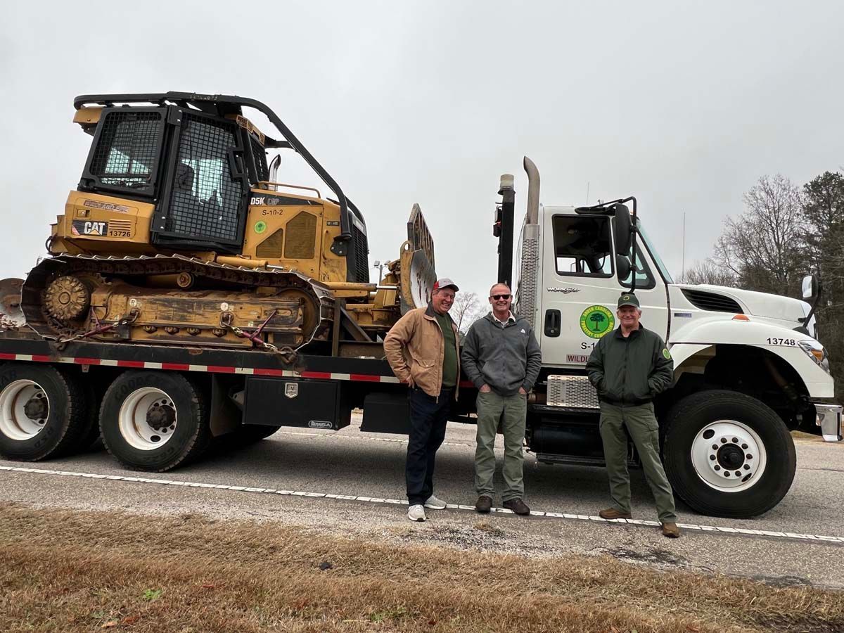 Three men posing beside a white truck hauling a yellow bulldozer on a flatbed trailer.