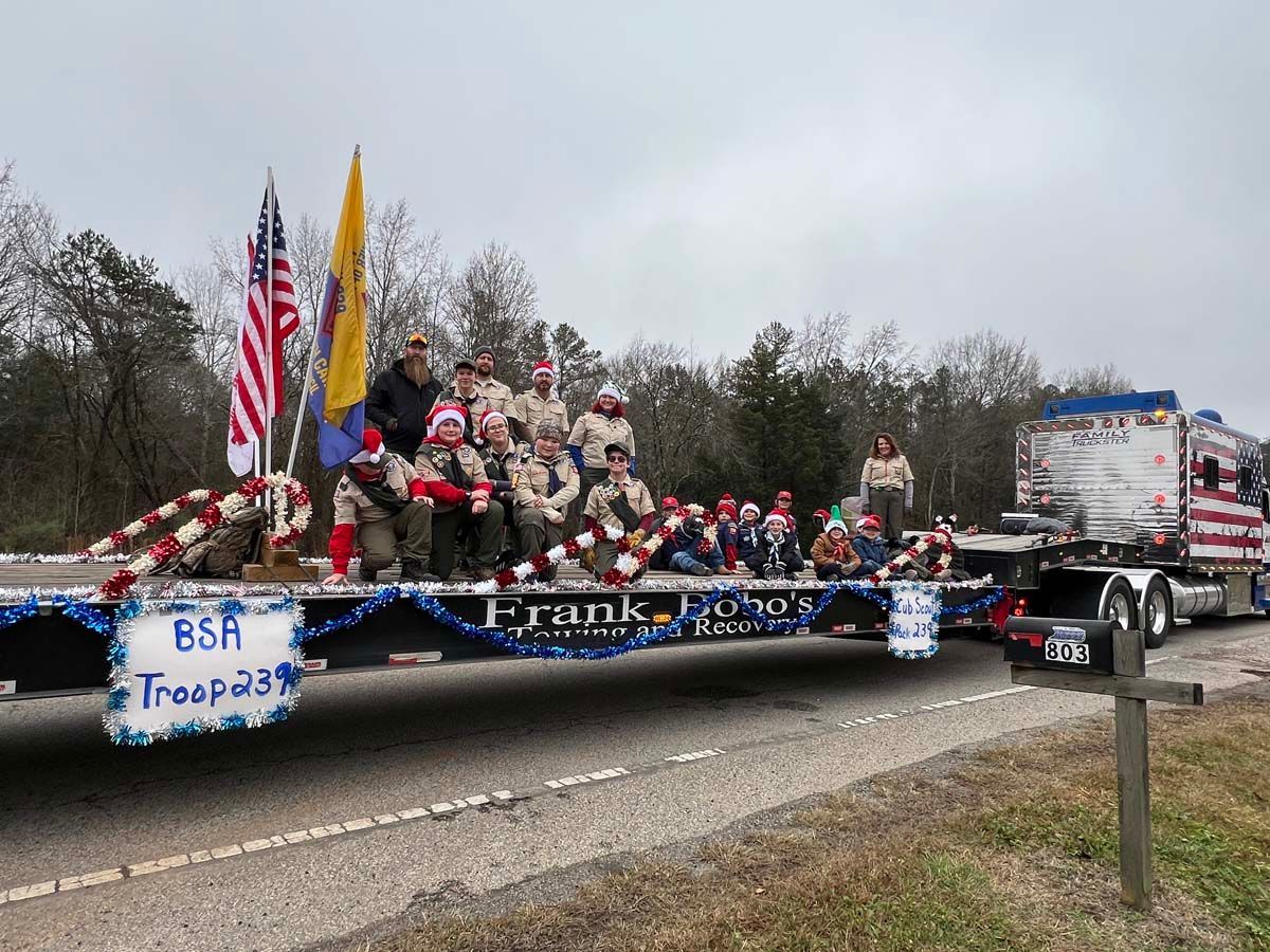 Decorated parade float with flags, people, and “Frank” banner, towing a truck along a road under cloudy skies