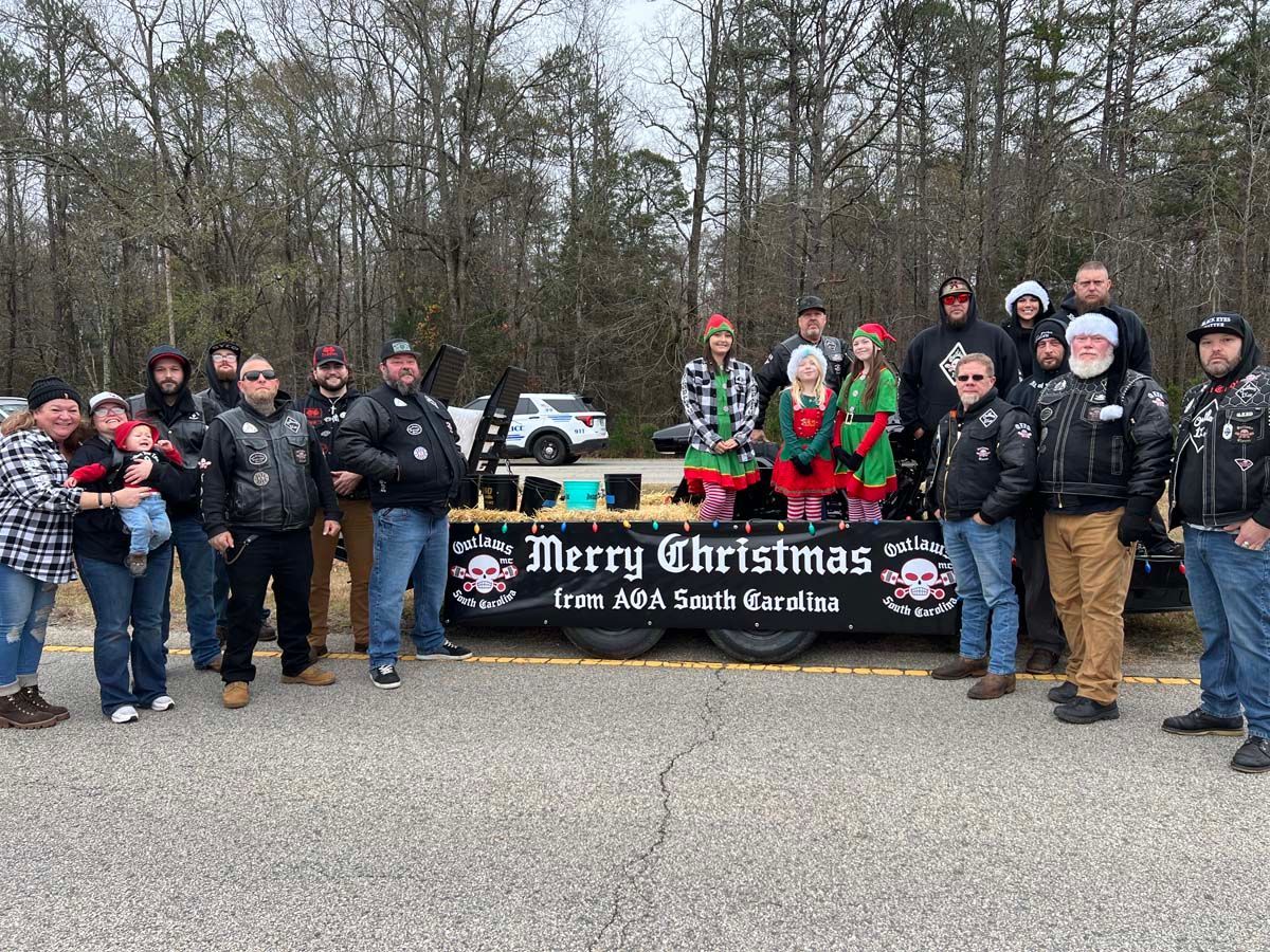 Group of people posing outdoors behind a “Merry Christmas” banner in front of trees and parked cars.