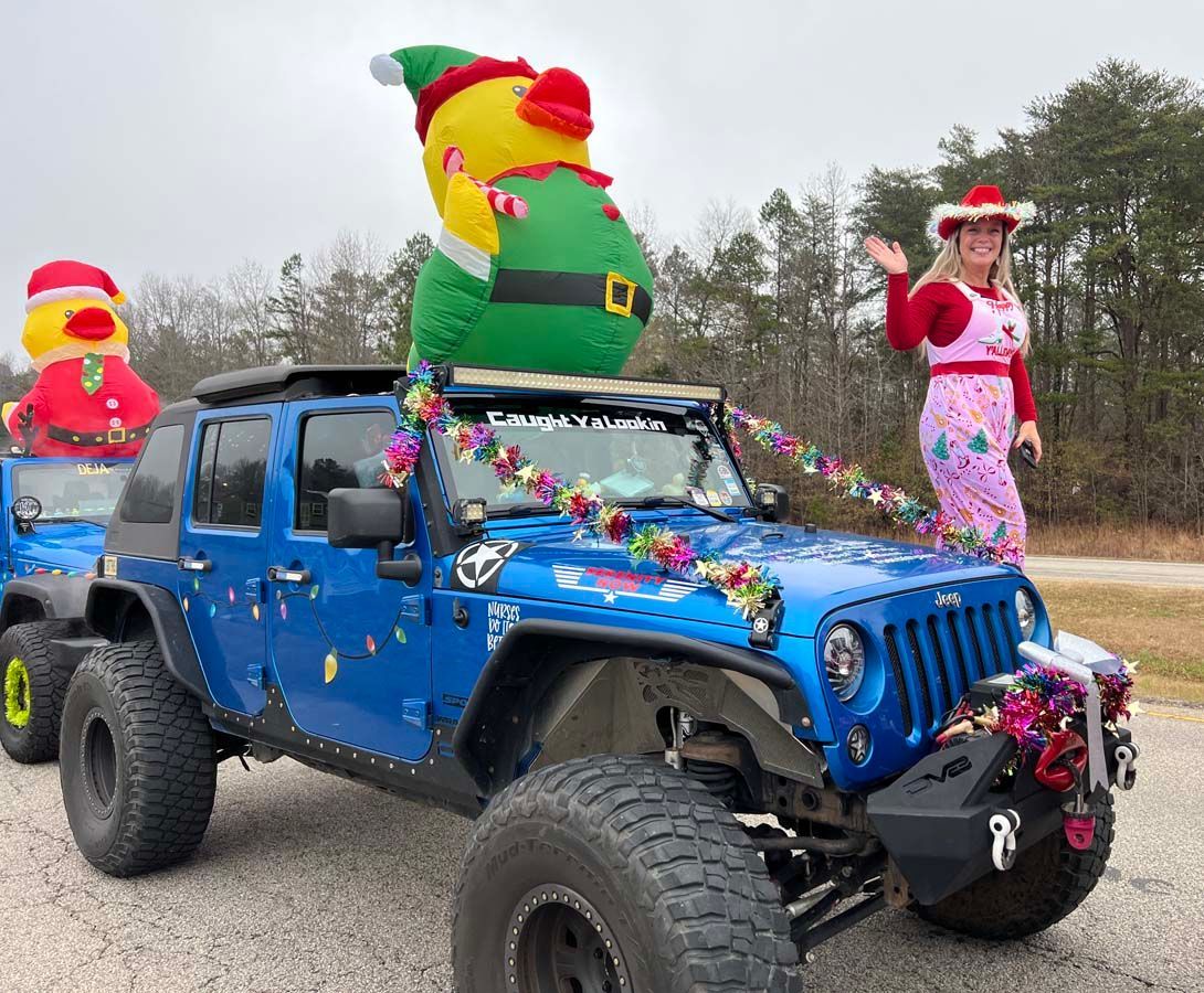 Blue Jeep with Christmas decorations and inflatable holiday figures on a road; a woman stands beside it.