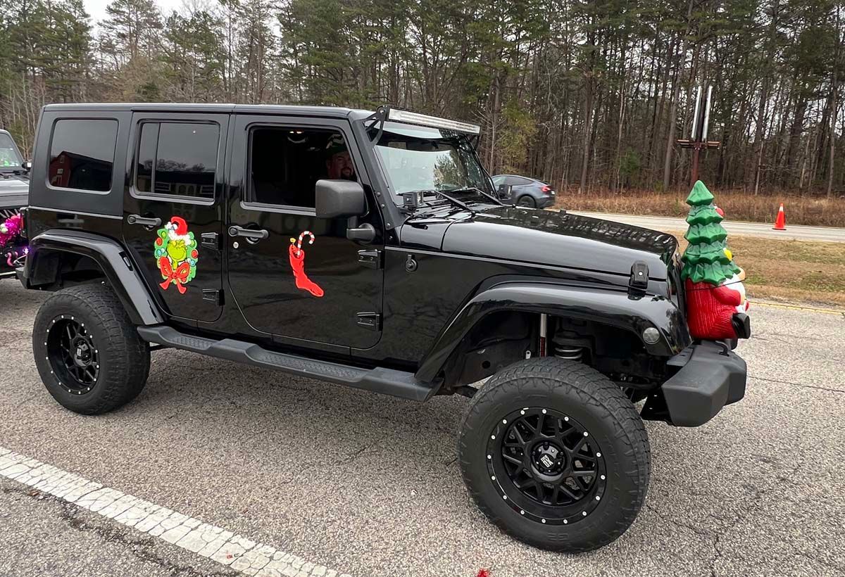 Black Jeep Wrangler decorated for Christmas, parked on a road beside a wooded area.