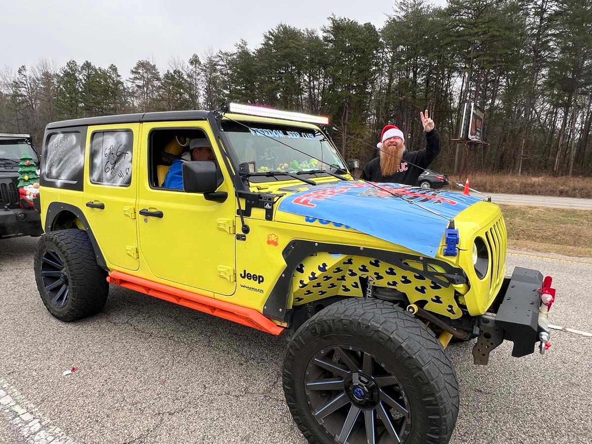 Bright yellow lifted Jeep with blue hood on a road, driver inside and a person posing through the roof.