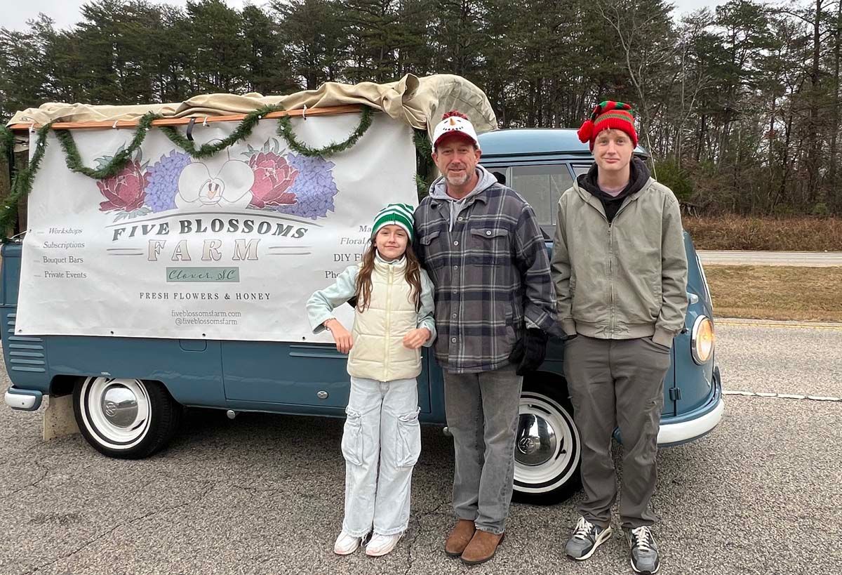 Three people stand in front of a decorated vintage blue van with a flower-covered sign.
