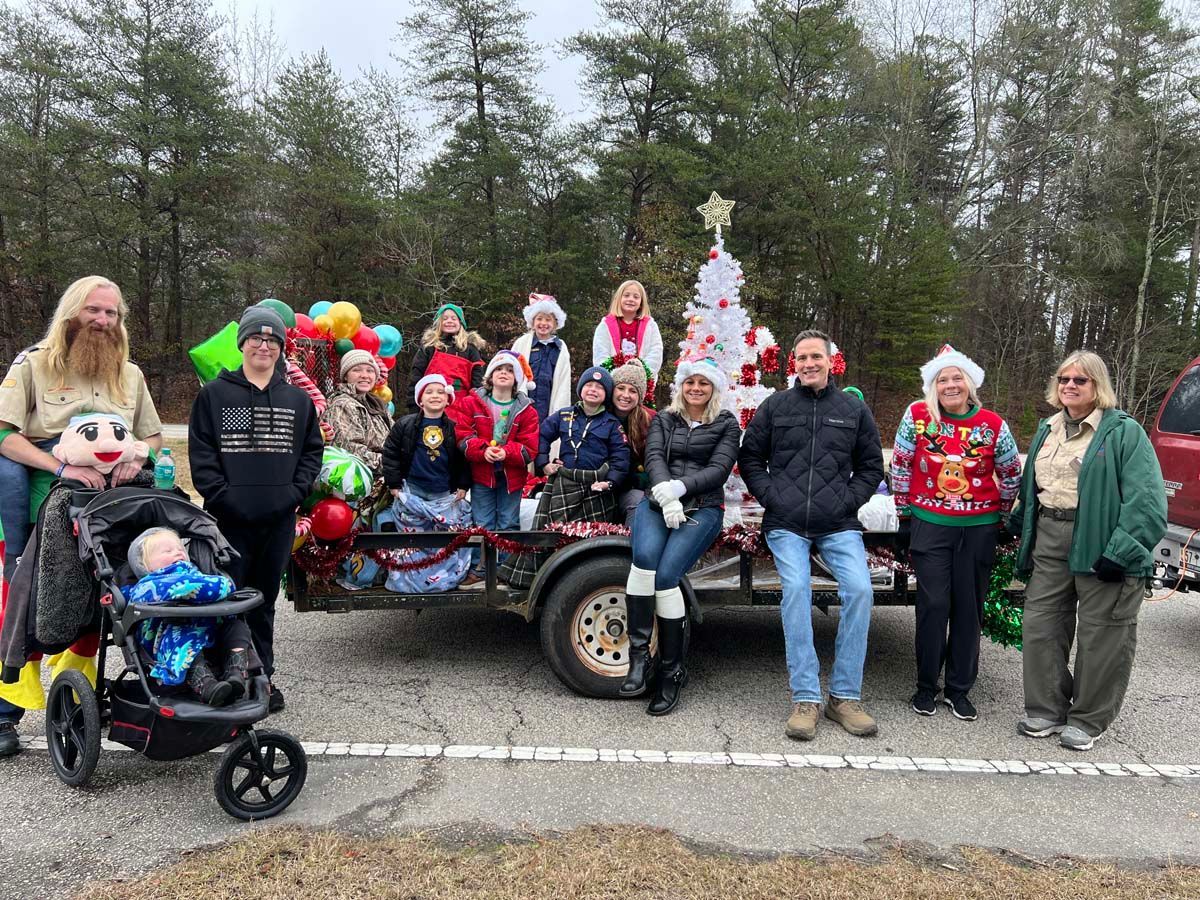 Group of people in festive hats gathered around a decorated trailer with a Christmas tree in a wooded area