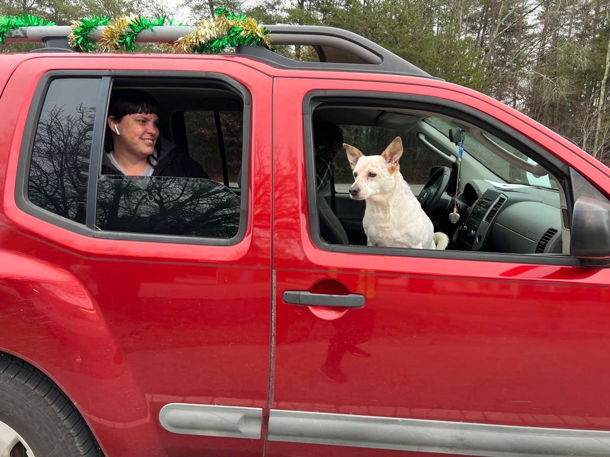 Red SUV with a white dog in the front seat and a smiling driver, outdoors with trees in the background