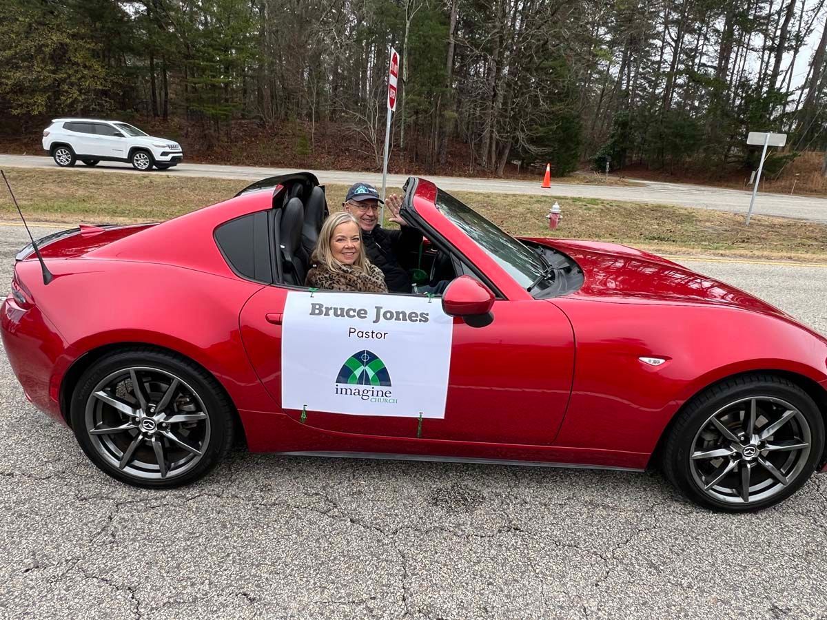 Red sports car with a “Mason Jones” campaign sign and two people inside on a roadside