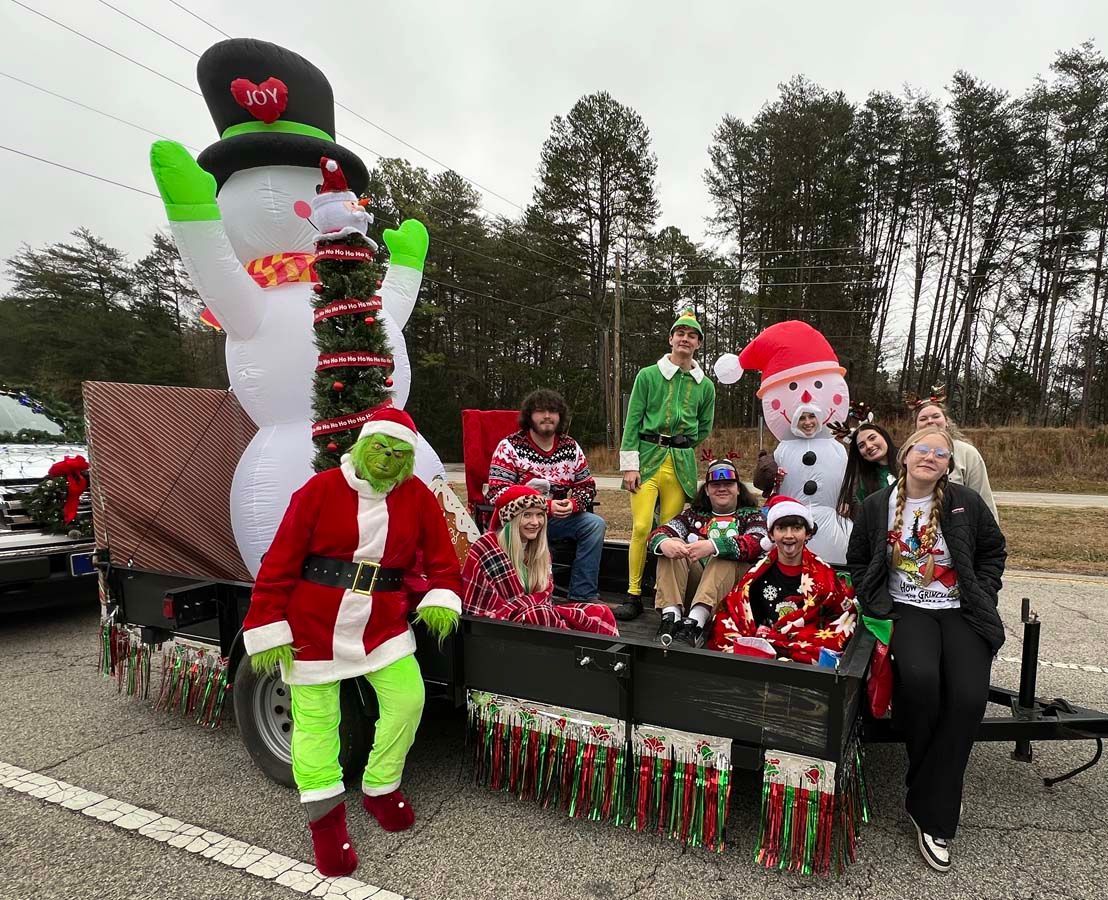 People posing on a festive parade float with oversized holiday decorations and Christmas costumes