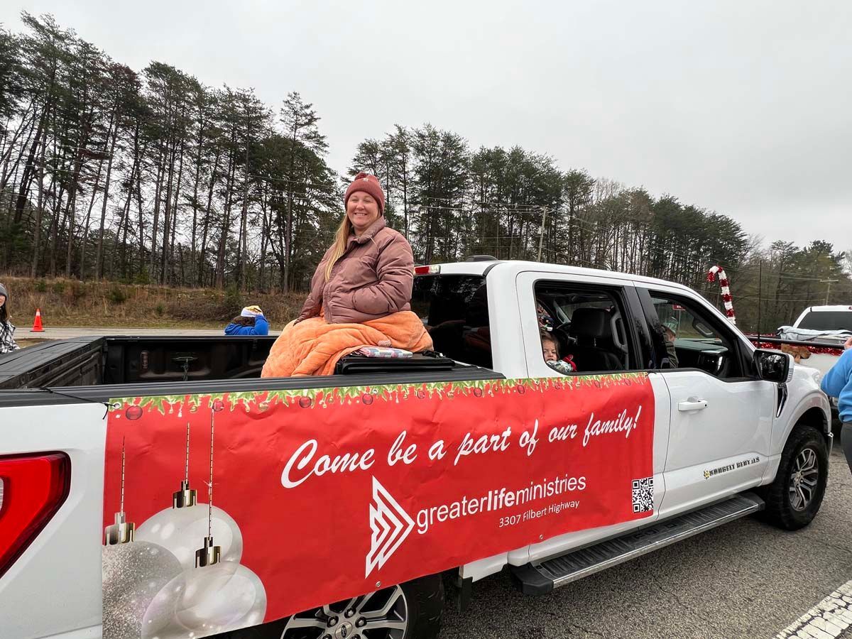 Person sitting in a white pickup truck with a red holiday banner on the side at a parade