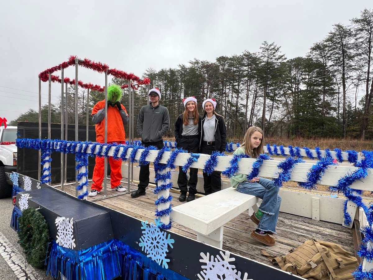 Decorated parade float with blue-and-white garlands and five people posing outdoors on a cloudy day