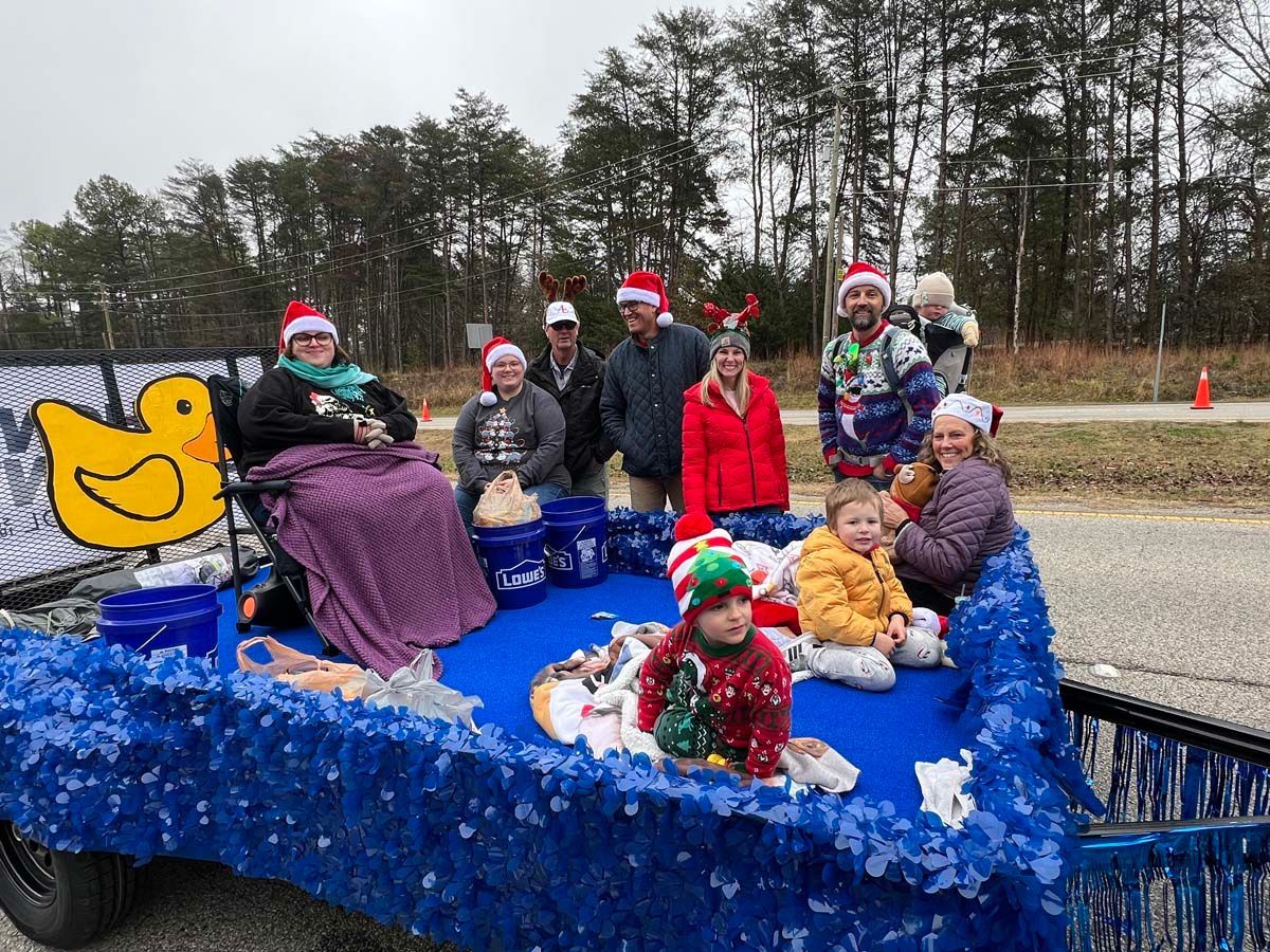 Holiday parade float with people in Santa hats on a blue, snow-covered cart with a yellow duck sign