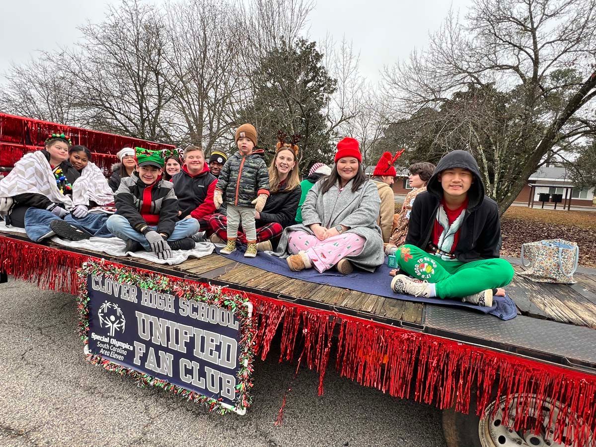 People seated on a decorated parade float with a “Hometown Fly Club” sign, outdoors on a cloudy day