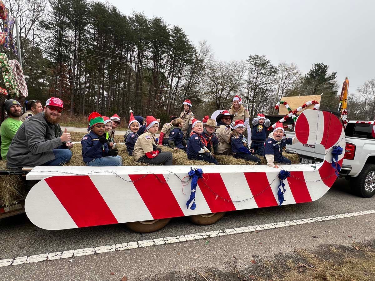 People in Santa hats ride on a parade float with red-and-white striped barriers.