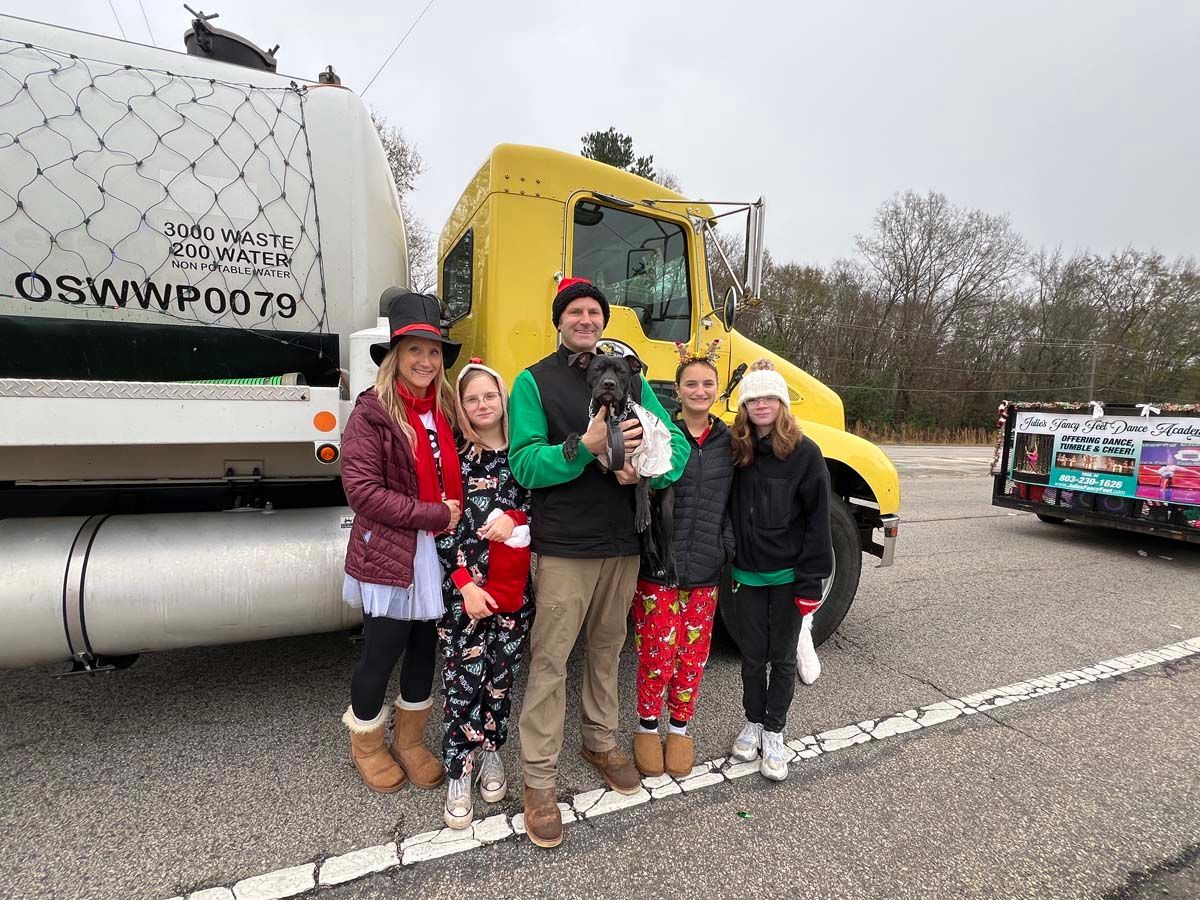 Group posing by a yellow truck and white tanker in a parking lot, wearing festive holiday clothing