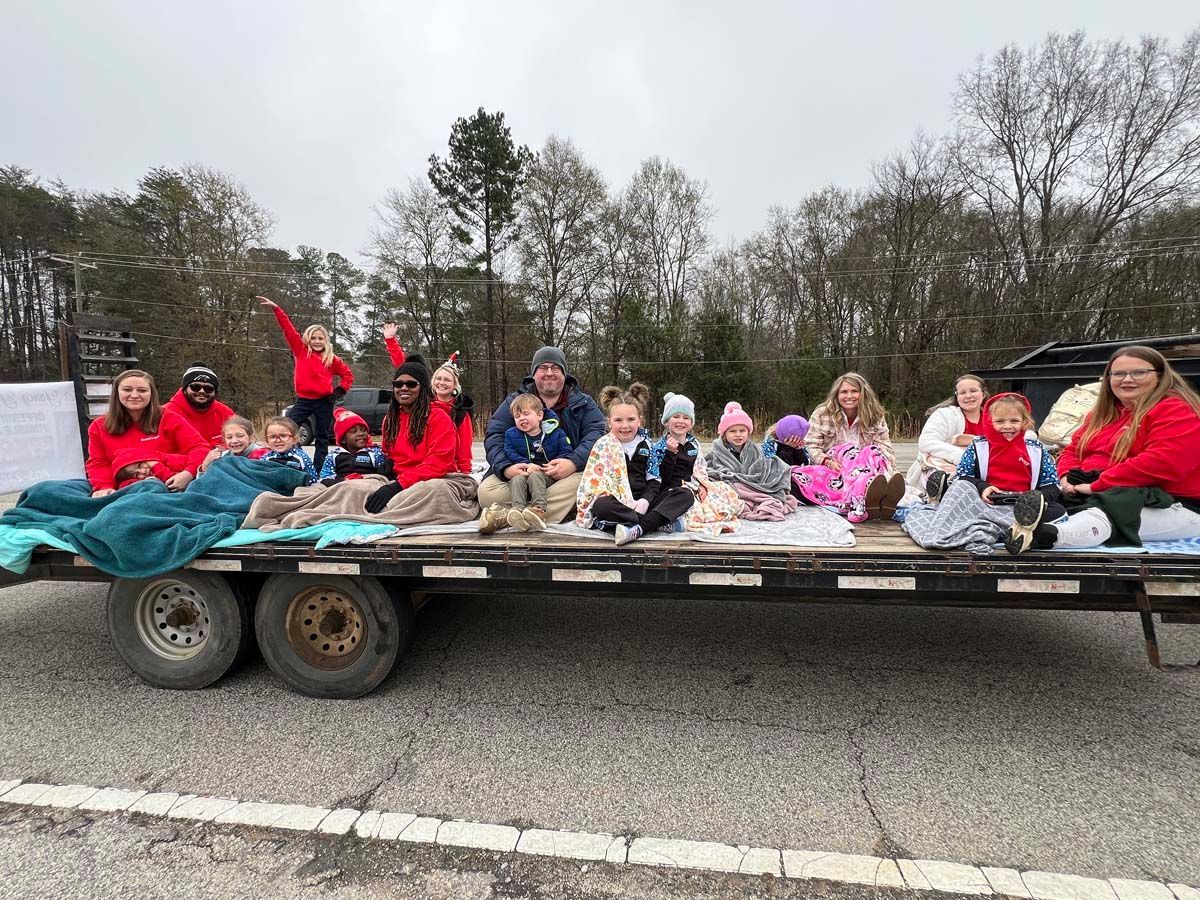Group of people sitting on a trailer, waving and smiling outdoors in a roadside parade scene
