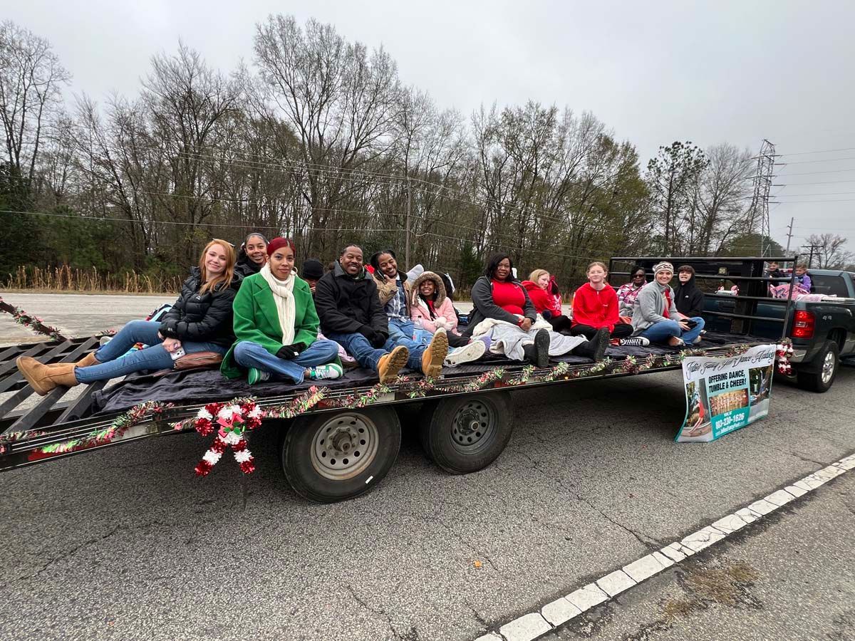 People seated on a decorated trailer being towed along a road, with trees and overcast sky in the background