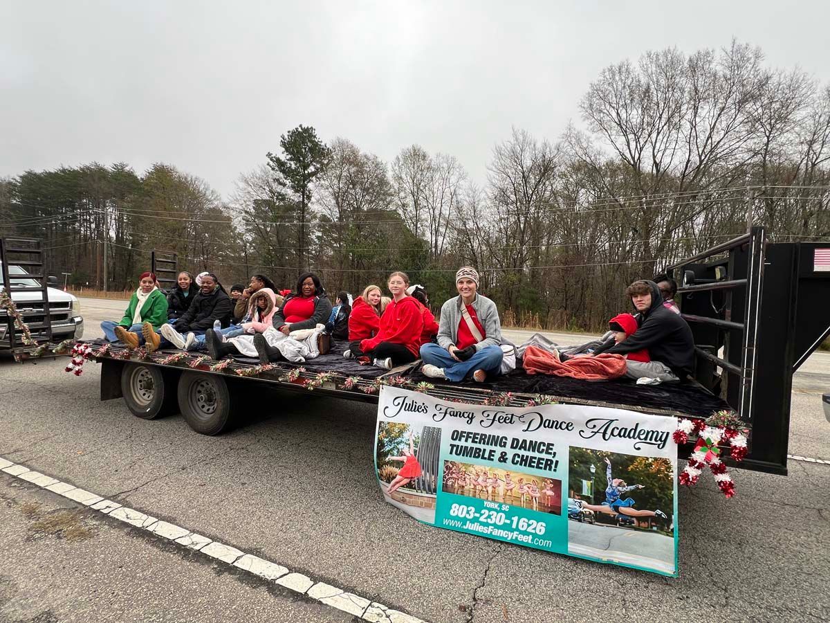 People sitting on a decorated parade float with a holiday banner on a road under cloudy skies