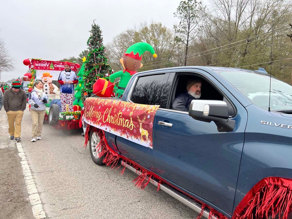 Blue pickup truck with red holiday decorations in a Christmas parade, driver visible, floats behind
