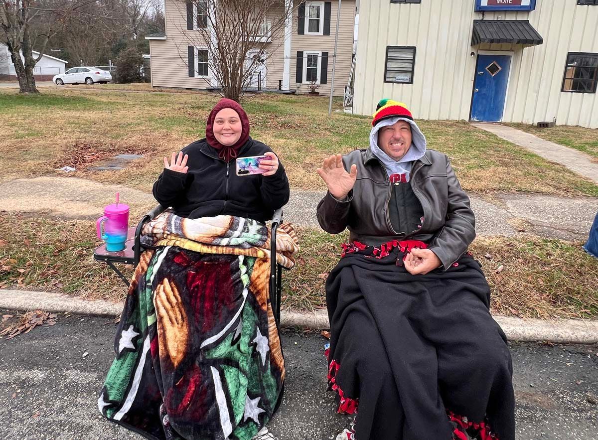 Two people seated outdoors in front of houses, smiling and waving, wrapped in blankets on a chilly day