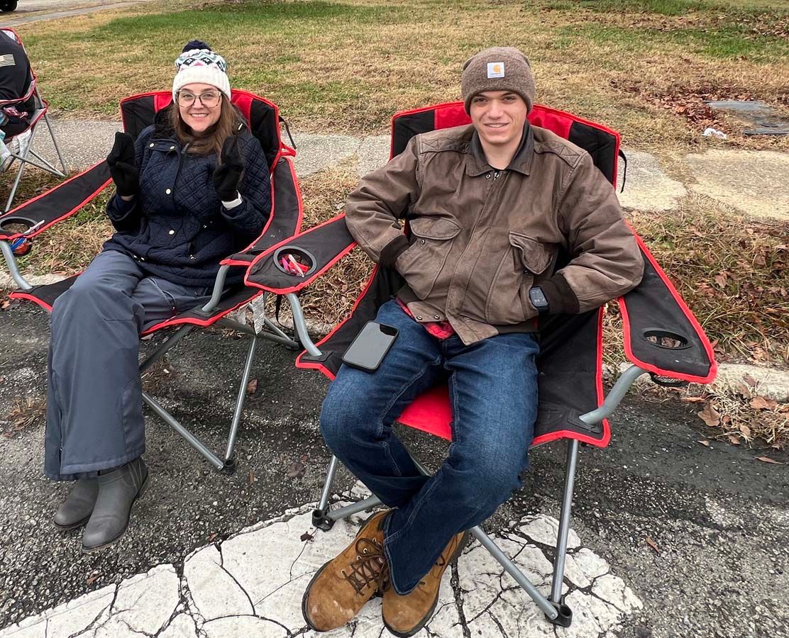 Two people sitting in red-and-black camp chairs outdoors on a paved roadside area.