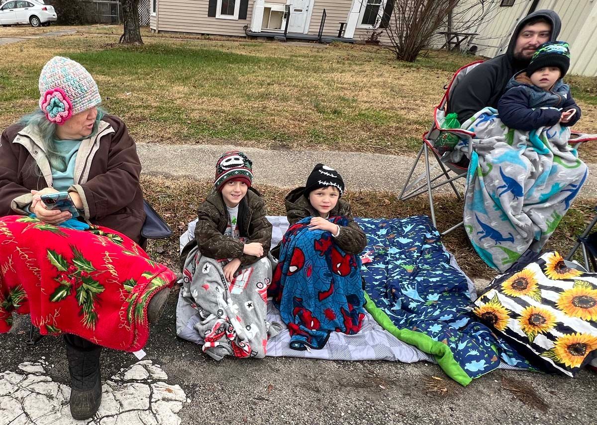 Several children and an adult sit outdoors on blankets, holding colorful quilt squares.