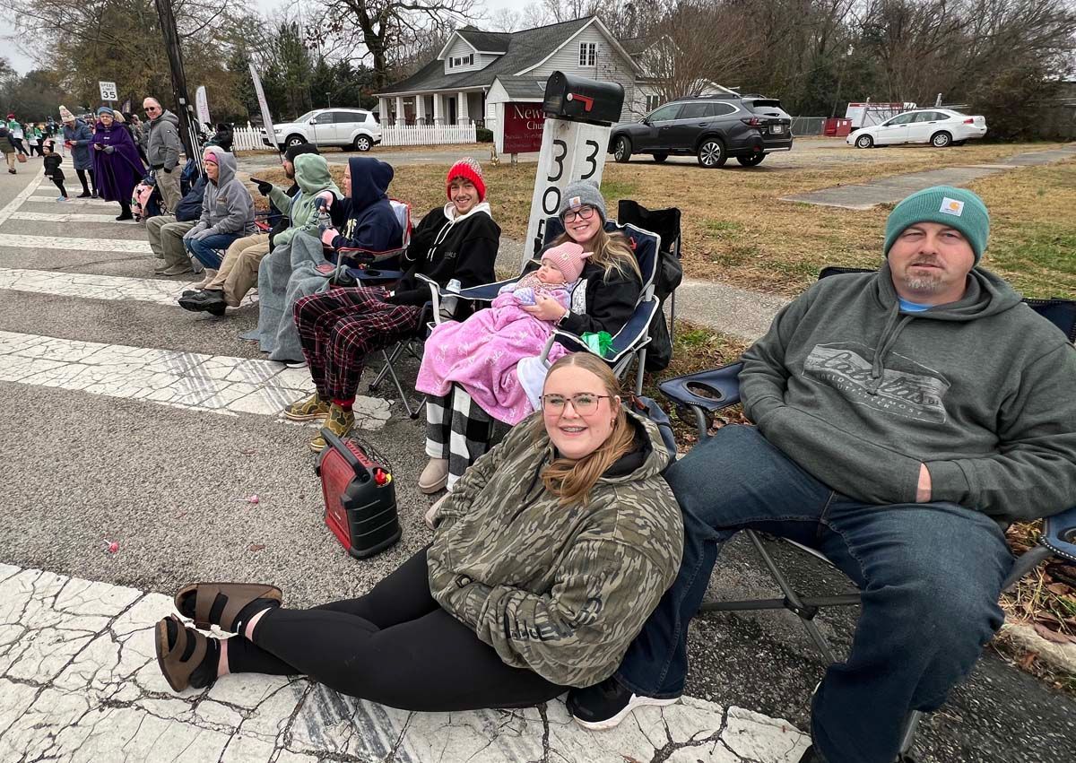 People seated in folding chairs along a street, bundled in winter clothes during a parade.