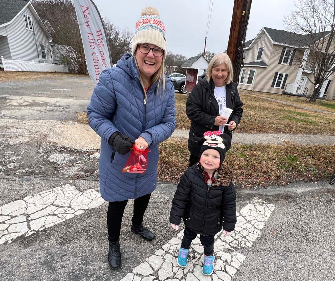 Three people in winter coats smiling outdoors on a sidewalk, one holding a red bag