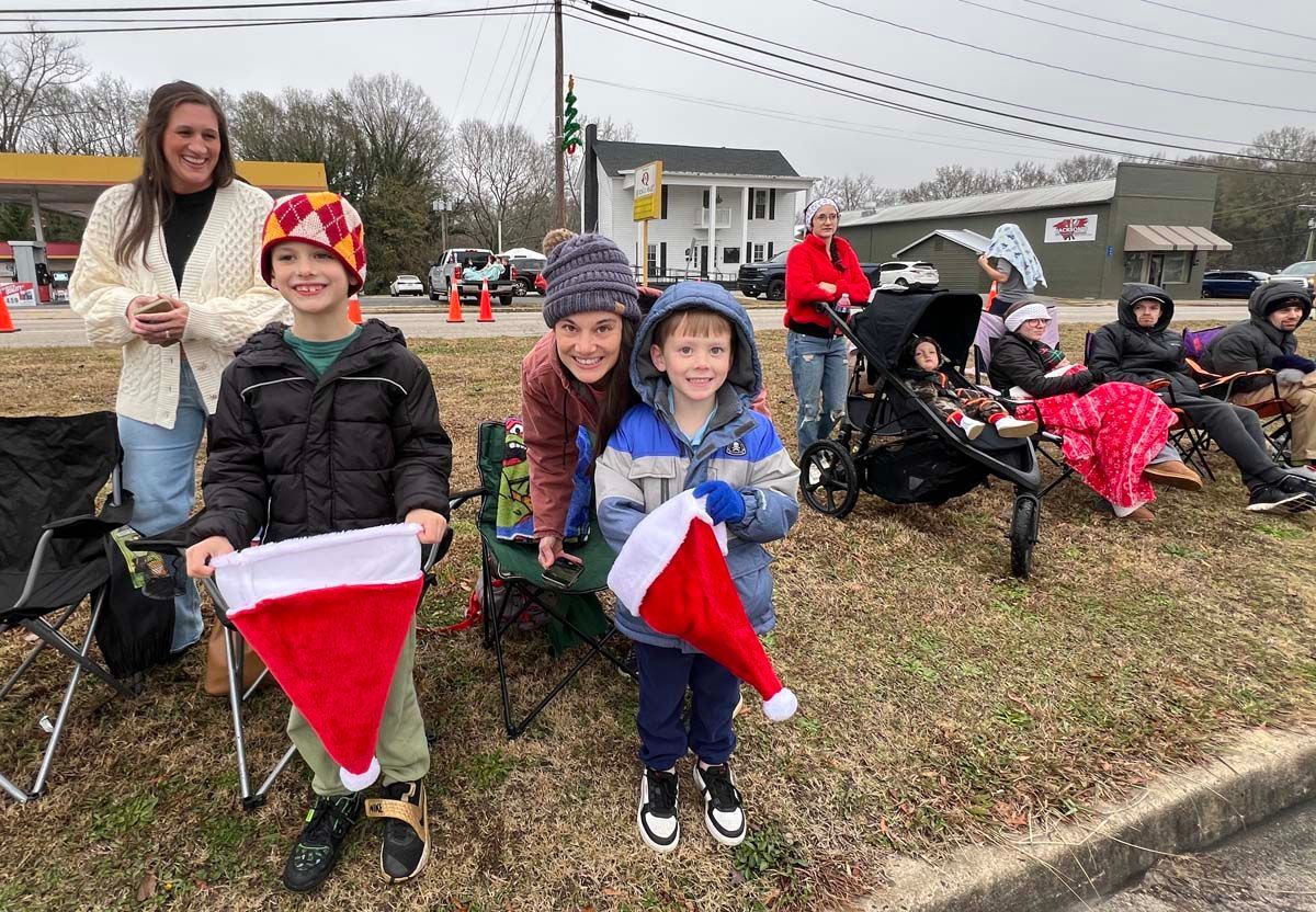 Children holding Santa hats at a roadside holiday parade, with spectators and a stroller in the background