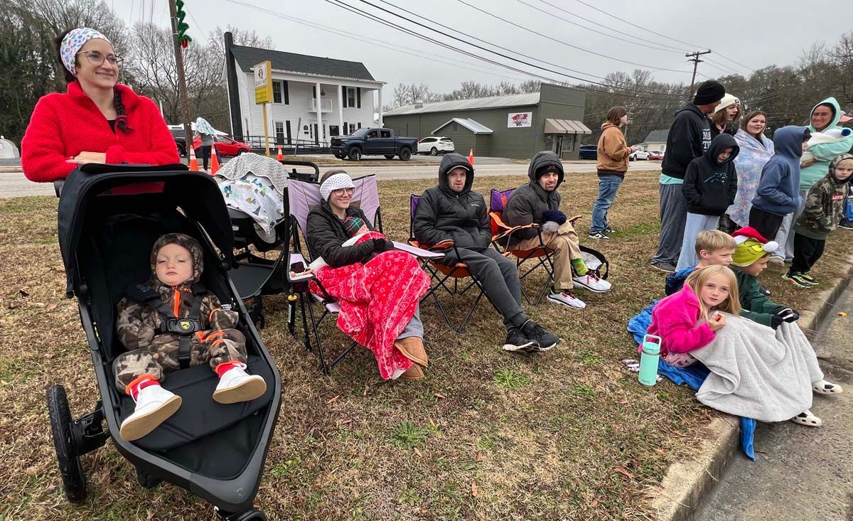 People seated outdoors along a curb, with strollers and bundled-up children at a roadside gathering