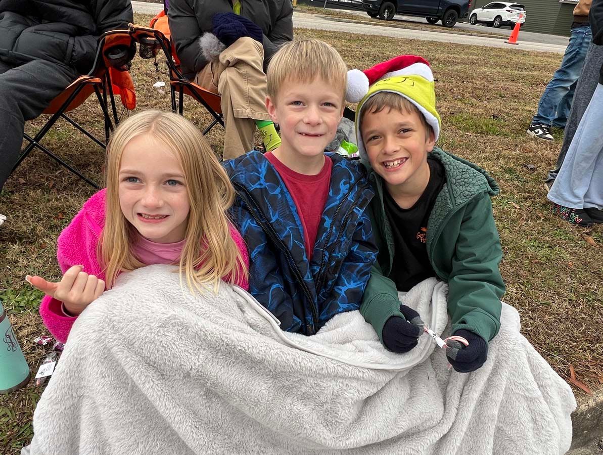 Three children smiling under a white blanket outdoors at a chilly event