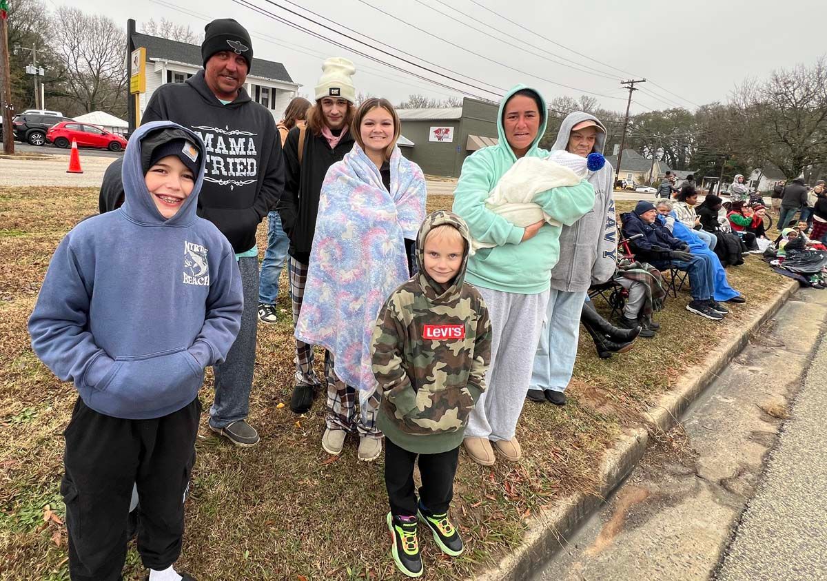 Family in winter coats standing along a curb, watching a roadside parade in cold weather.