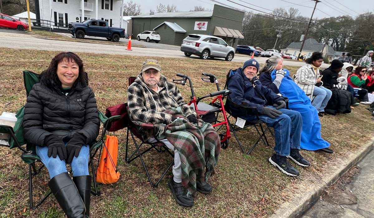 People seated on benches along a roadside, bundled in winter coats and blankets on an overcast day.